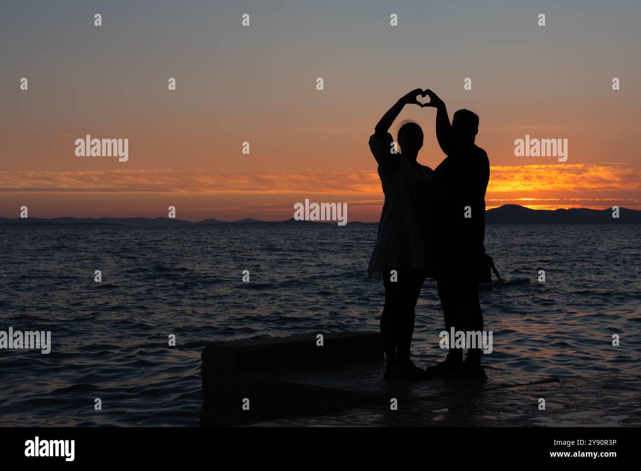 Silhouette di una coppia che crea una forma a cuore con le mani al tramonto in riva al mare. Foto Stock