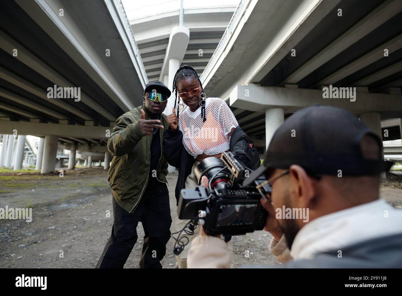 Diversi gruppi di persone che partecipano alla realizzazione di video su scene urbane sotto ponti autostradali sopraelevati, con un membro della troupe che si concentra sui soggetti e sull'ambiente circostante Foto Stock