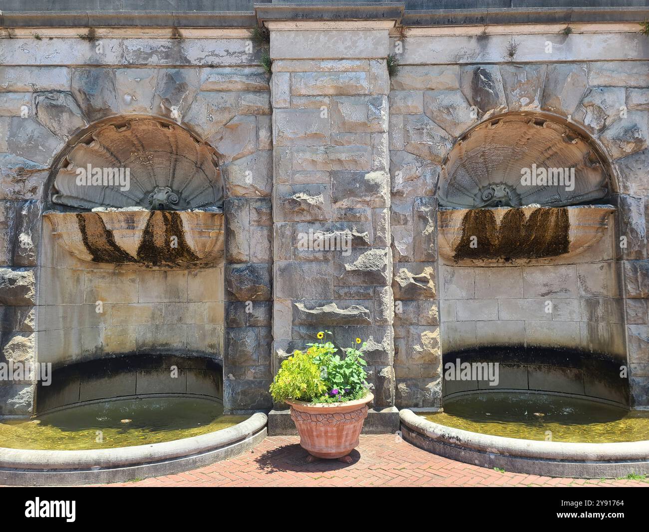 Un muro di pietra con due alcove ad arco, ciascuna contenente una piccola fontana. Una pianta in vaso con fiori è posta di fronte al muro su una superficie di mattoni. Foto Stock