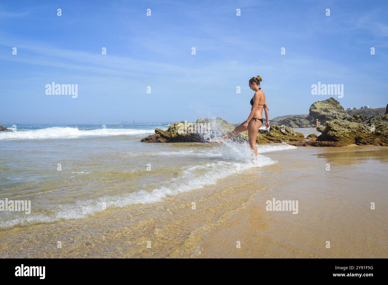 Una donna sulla spiaggia gode del mare mentre le onde si tuffano intorno ai suoi piedi. Il cielo è blu e soleggiato Foto Stock