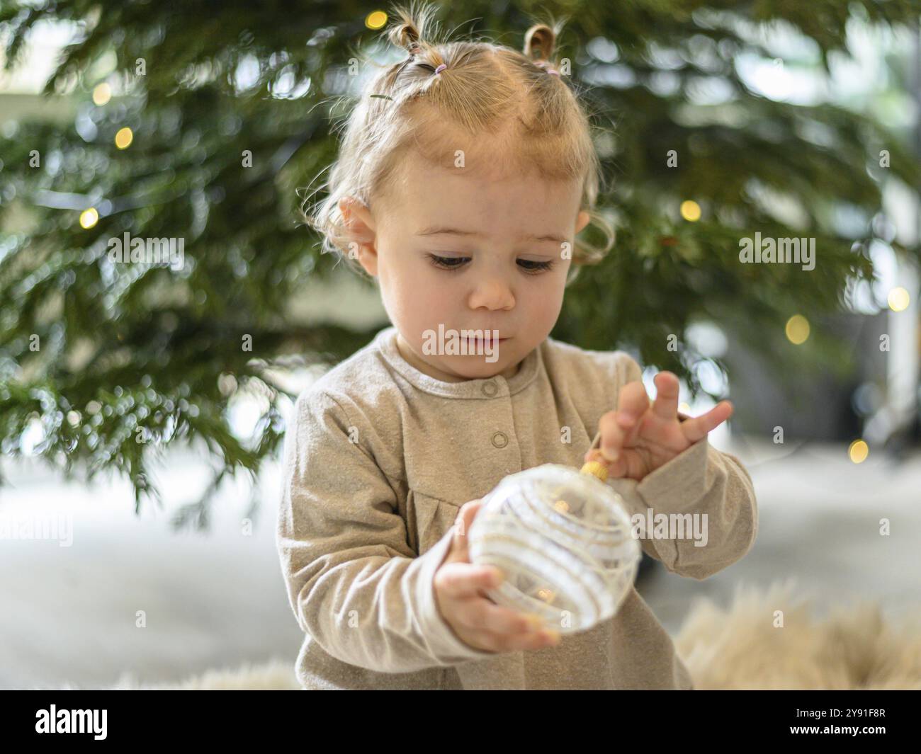 Il bambino ispeziona una palla trasparente sull'albero di Natale di fronte a un albero di Natale illuminato Foto Stock