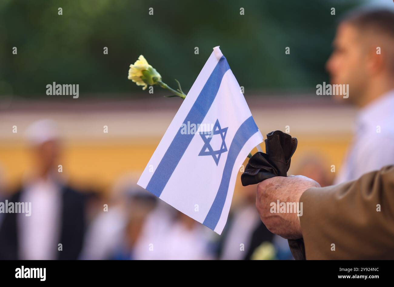 Dettagli con le mani di un uomo che regge la bandiera israeliana e un fiore durante una cerimonia commemorativa degli attacchi dell'ottobre 2023. Foto Stock