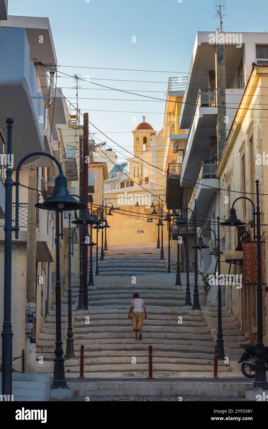 Via Grecia, vista posteriore al crepuscolo di una donna che cammina su una scala situata in una strada nel quartiere della città vecchia di Sitia, nel nord-est di Creta, in Grecia. Foto Stock