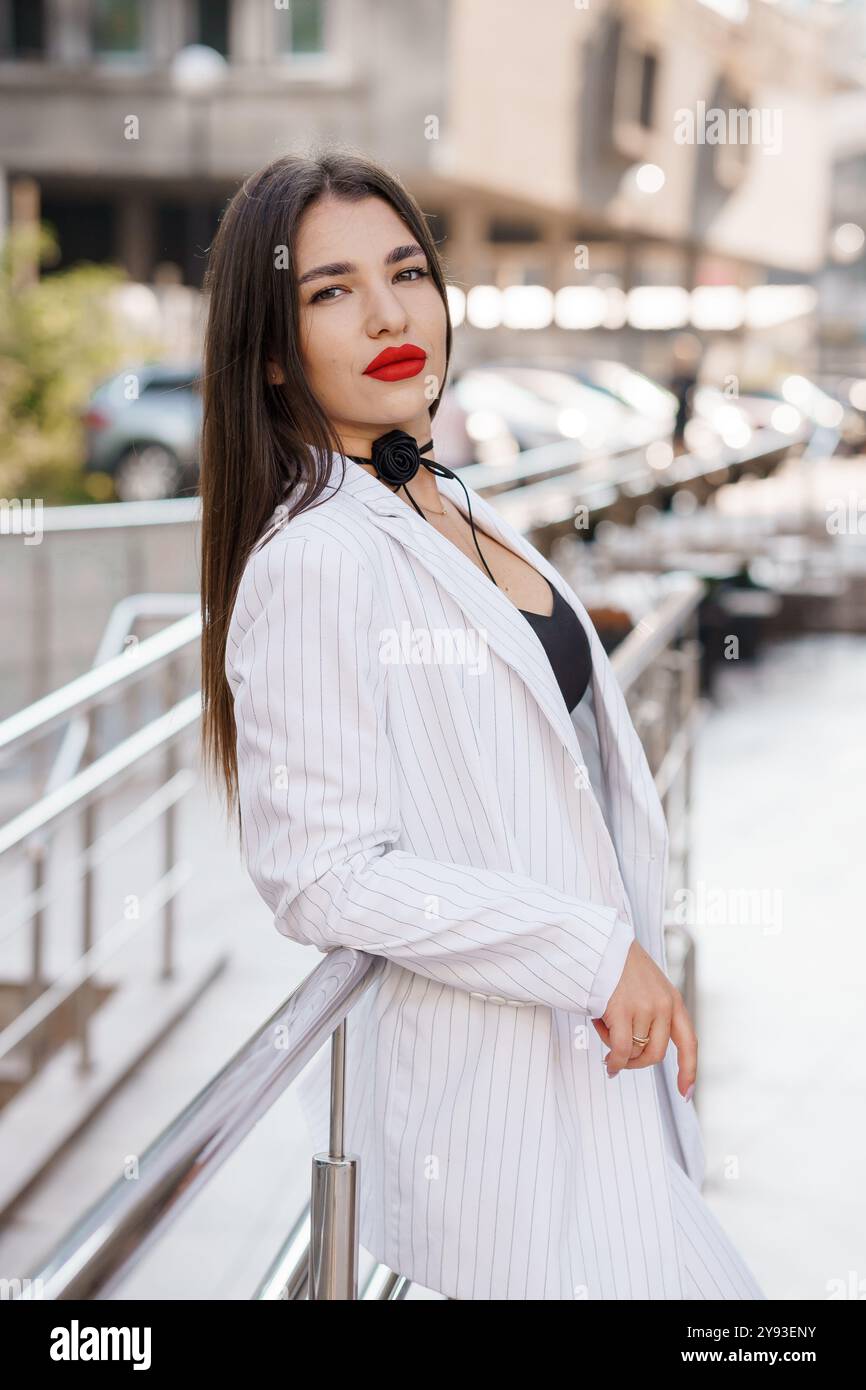 Donna elegante che posa all'aperto con sicurezza in un elegante abito a righe con un'audace labbro rosso e un accessorio floreale. Foto Stock