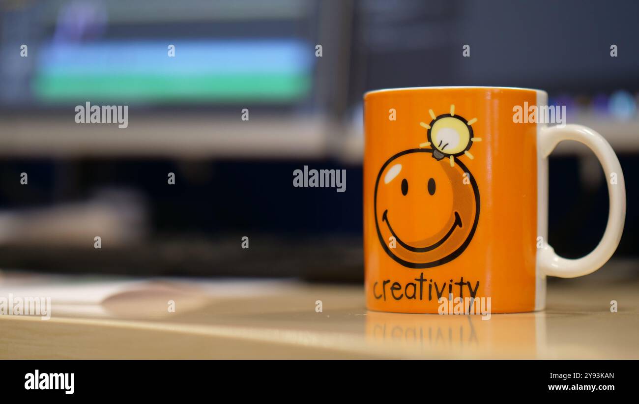 Un'allegra tazza arancione con un volto sorridente e la parola "creatività" scritta su di essa, caratterizzata da un design a lampadina che rende l'ufficio sfocato sullo sfondo Foto Stock Un'allegra tazza arancione con un volto sorridente e la parola "creatività" scritta su di essa, caratterizzata da un design a lampadina che rende l'ufficio sfocato sullo sfondo Foto Stock