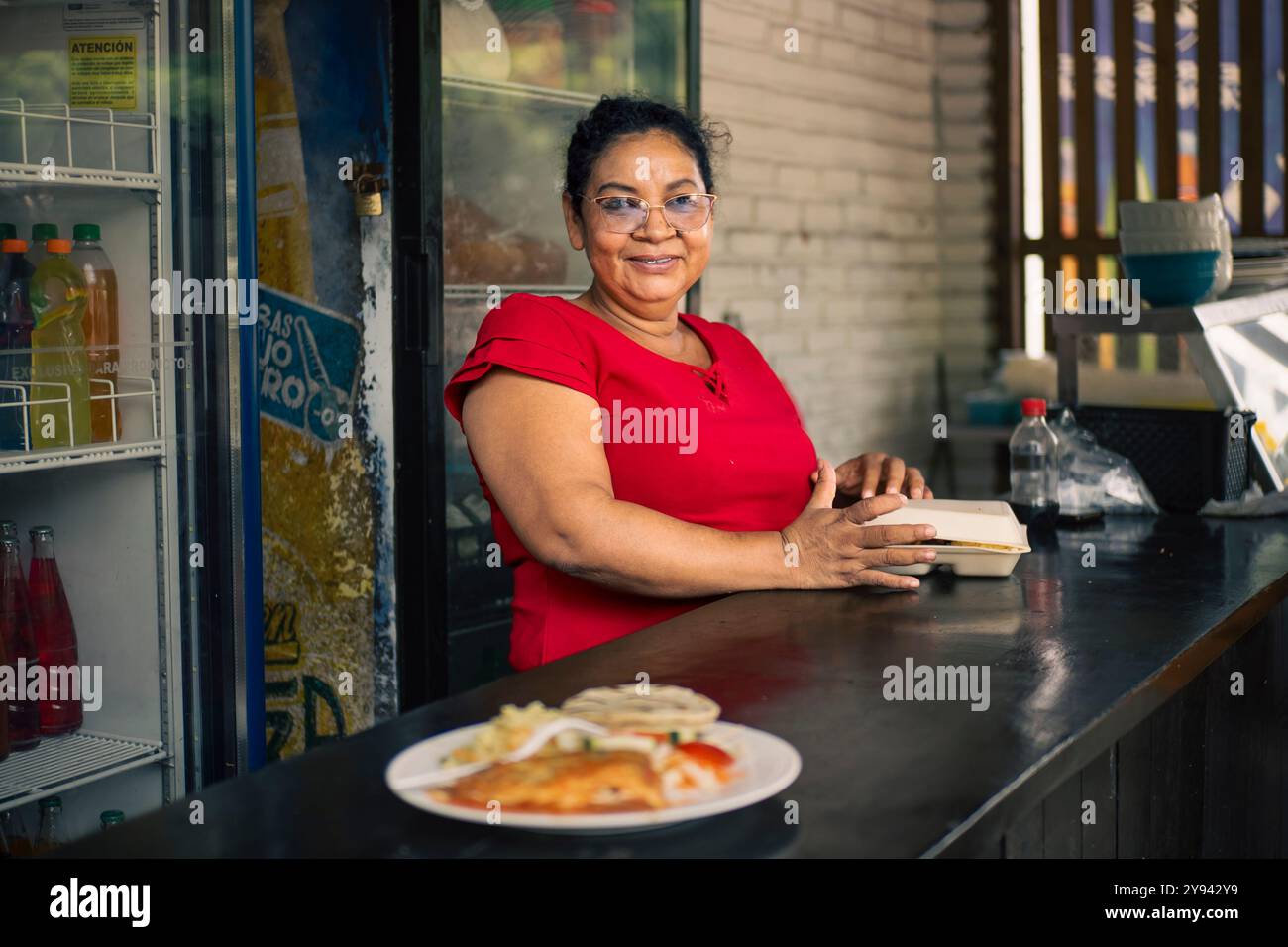 Una allegra donna salvadoregna serve cibo di strada tradizionale dal suo chiosco, mettendo in risalto la cultura e la cucina locale in El Salvador Foto Stock