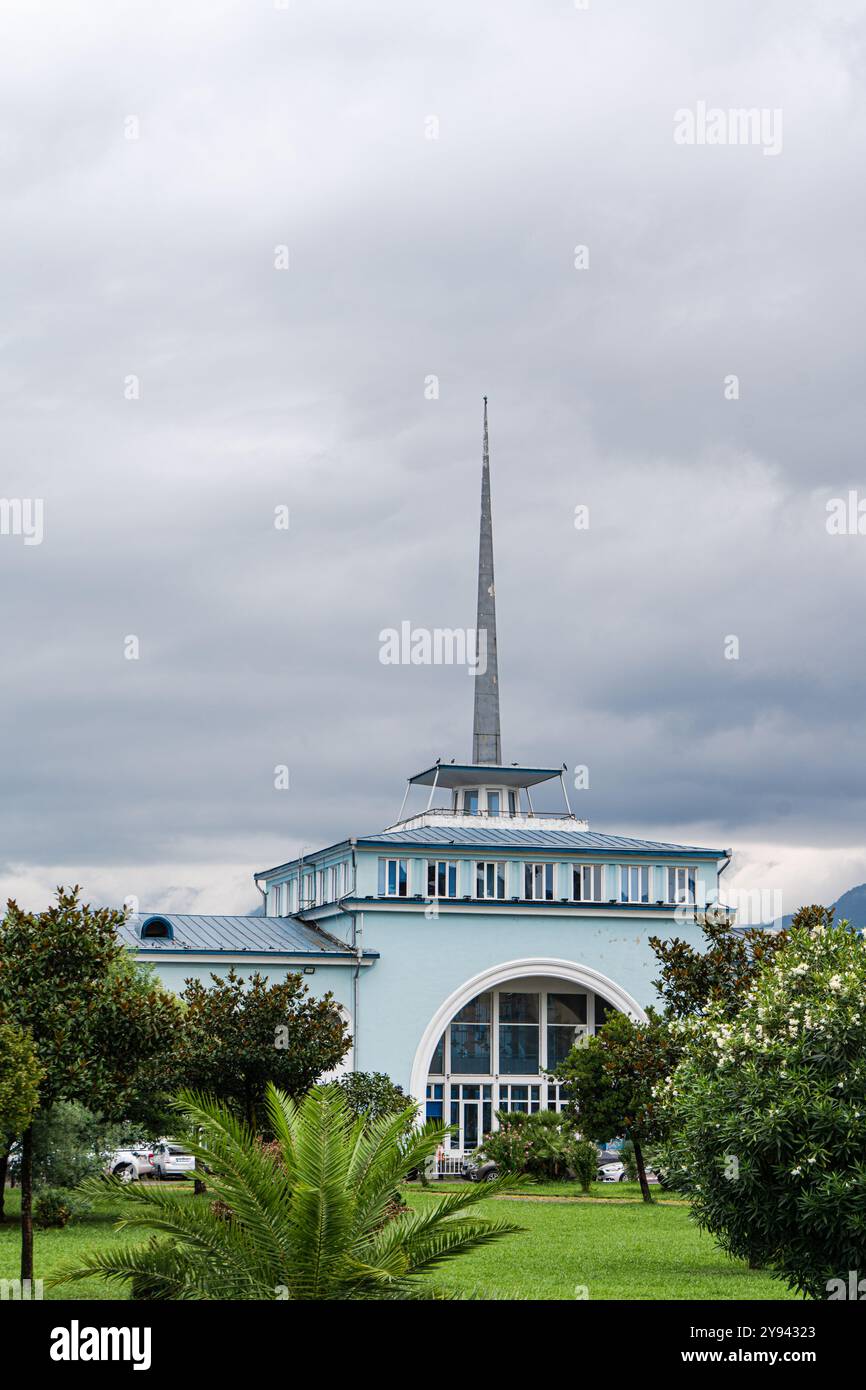 Un'immagine suggestiva che mostra un edificio di stile moderno con una prominente guglia, incorniciata da lussureggiante vegetazione e un cielo nuvoloso, raffigurante una architettura unica Foto Stock