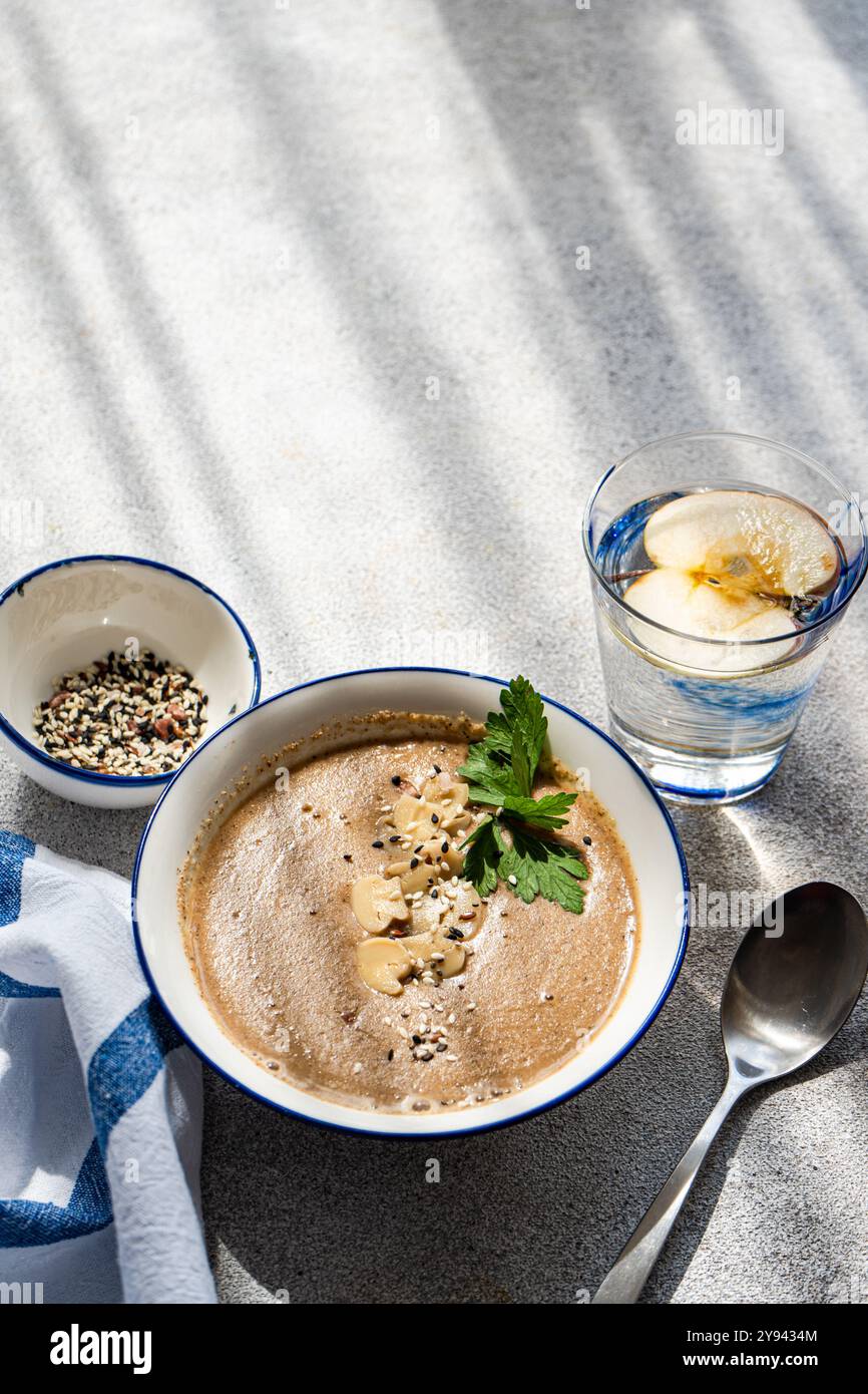 Vista dall'alto di una sana cena con zuppa di crema di funghi guarnita con semi ed erbe aromatiche, accanto a un bicchiere d'acqua infuso con mela biologica Foto Stock