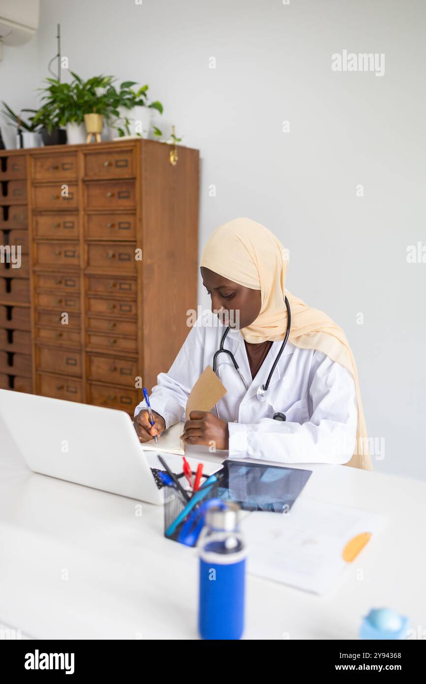 Un medico professionista di colore che indossa un hijab si concentra sulla scrittura di appunti mentre si siede alla sua scrivania in un ambiente d'ufficio moderno e ben illuminato, ref Foto Stock