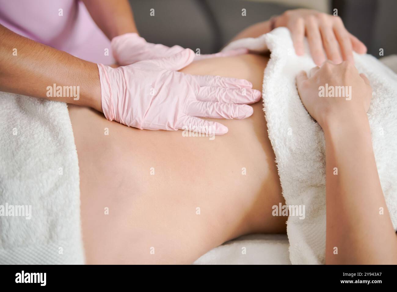 Massaggiatrice professionale corta e irriconoscibile, con guanti rosa, offre un massaggio rilassante allo stomaco a una paziente sdraiata su un tavolo terapeutico. T Foto Stock