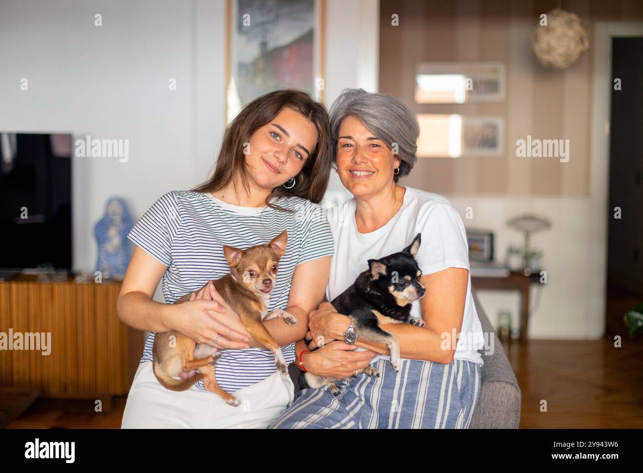 Madre matura felice e figlia adulta, sorridenti e con i loro piccoli cani Chihuahua, comodamente seduti in un ambiente accogliente Foto Stock