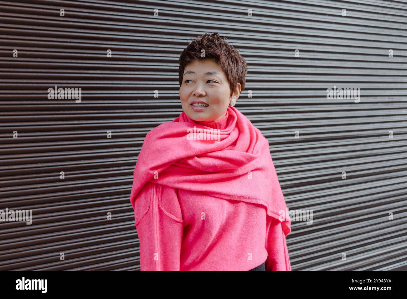 Una gioiosa donna asiatica di mezza età avvolta da una vivace sciarpa rosa si erge davanti a uno sfondo a righe scure, che irradia calore e stile in un ambiente informale all'aperto Foto Stock