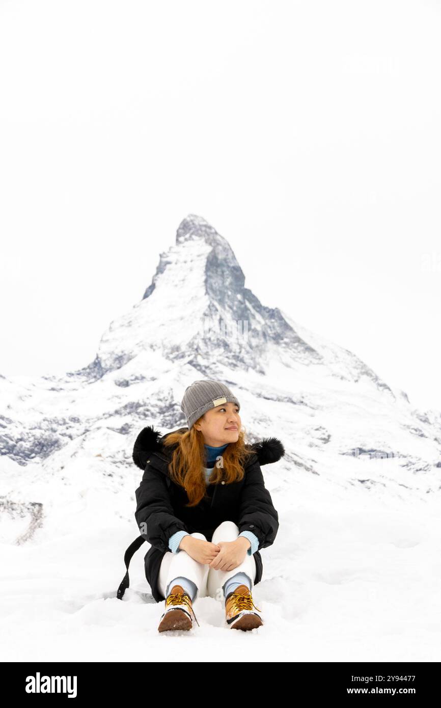Una giovane donna asiatica, che indossa una giacca invernale nera e un berretto grigio, si siede sulla neve. Ha uno sguardo attento con un maestoso opaco coperto di neve Foto Stock