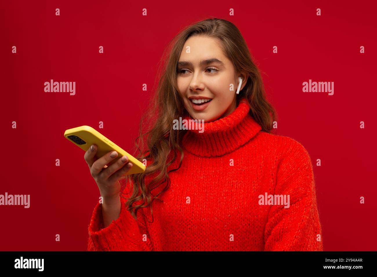 Giovane donna emotiva che ascolta la musica con gli auricolari wireless utilizzando uno smartphone moderno su uno sfondo rosso da studio Foto Stock