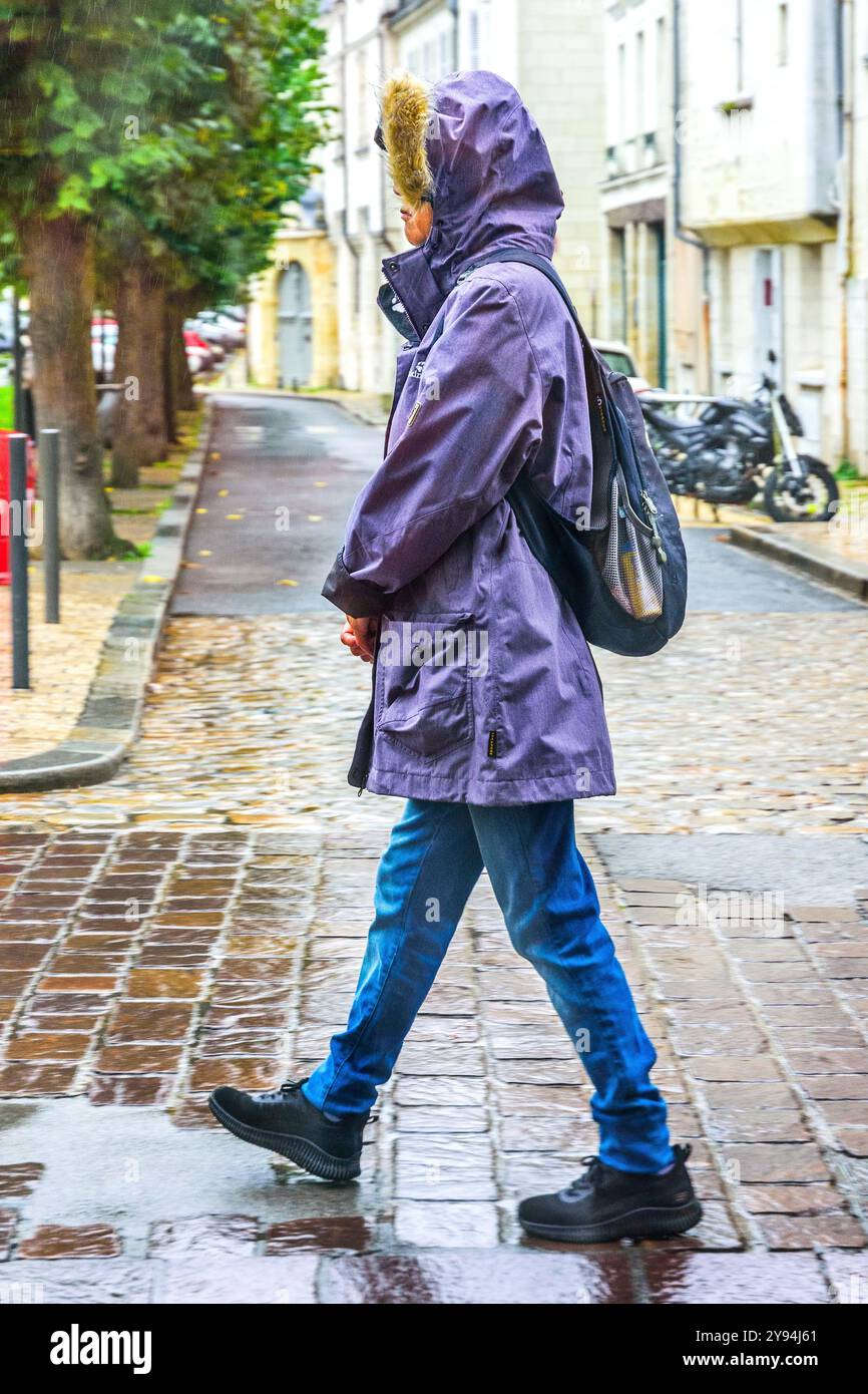 Donna con abbigliamento da pioggia che cammina lungo la strada della città sotto la pioggia battente senza ombrello - Tours, Indre-et-Loire (37), Francia. Foto Stock