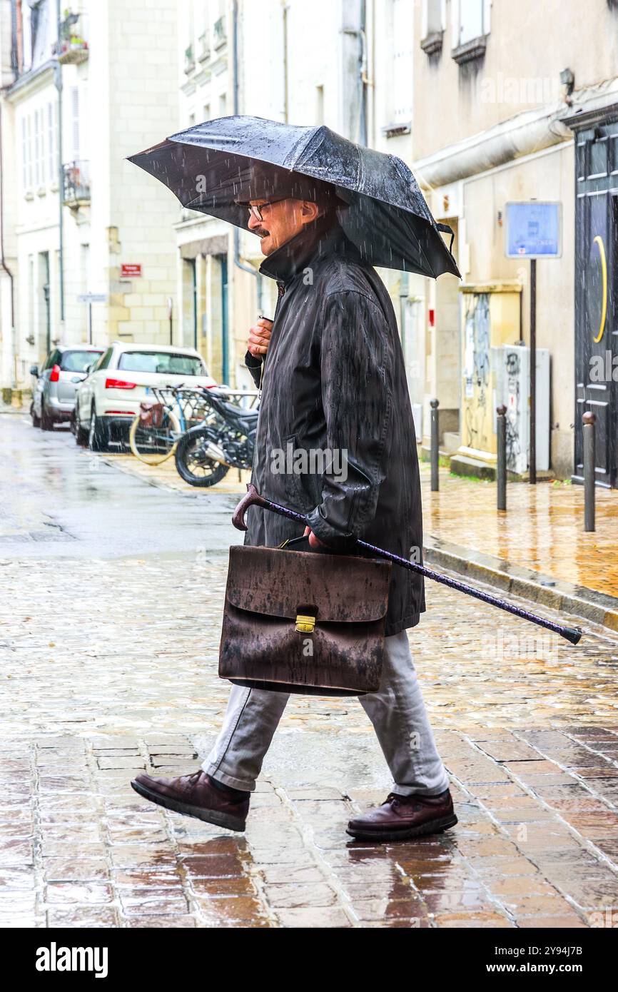 Uomo d'affari anziano con valigetta, bastone da passeggio e ombrello che cammina lungo la strada della città sotto la pioggia battente - Tours, Indre-et-Loire (37), Francia. Foto Stock