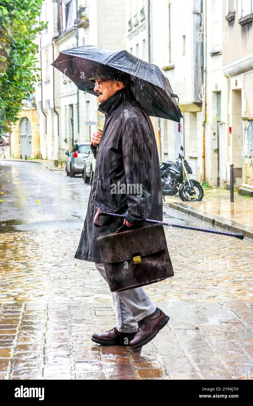 Uomo d'affari anziano con valigetta, bastone da passeggio e ombrello che cammina lungo la strada della città sotto la pioggia battente - Tours, Indre-et-Loire (37), Francia. Foto Stock