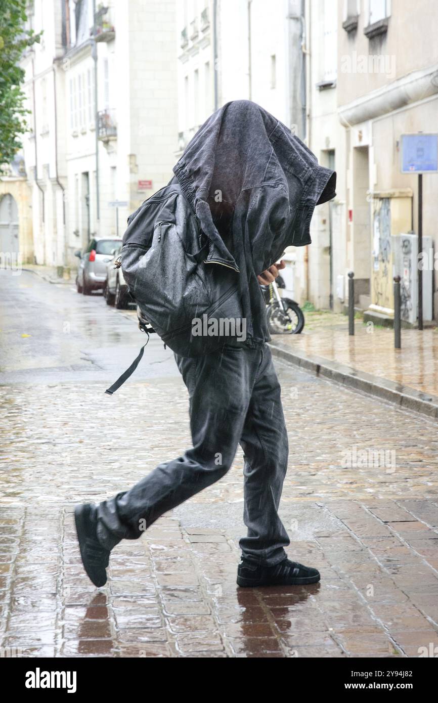 Uomo che cammina lungo la strada della città sotto la pioggia battente con giacca che copre testa e spalle - Tours, Indre-et-Loire (37), Francia. Foto Stock