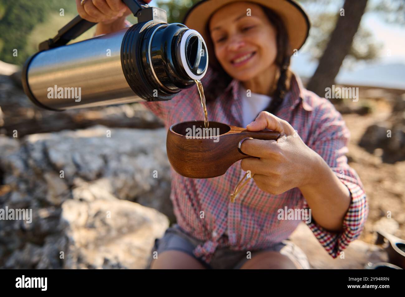 Una donna allegra versa una bevanda calda da un thermos in una tazza di legno mentre si gode la vita all'aperto. La luce del sole e l'ambiente naturale suggeriscono il relax a. Foto Stock