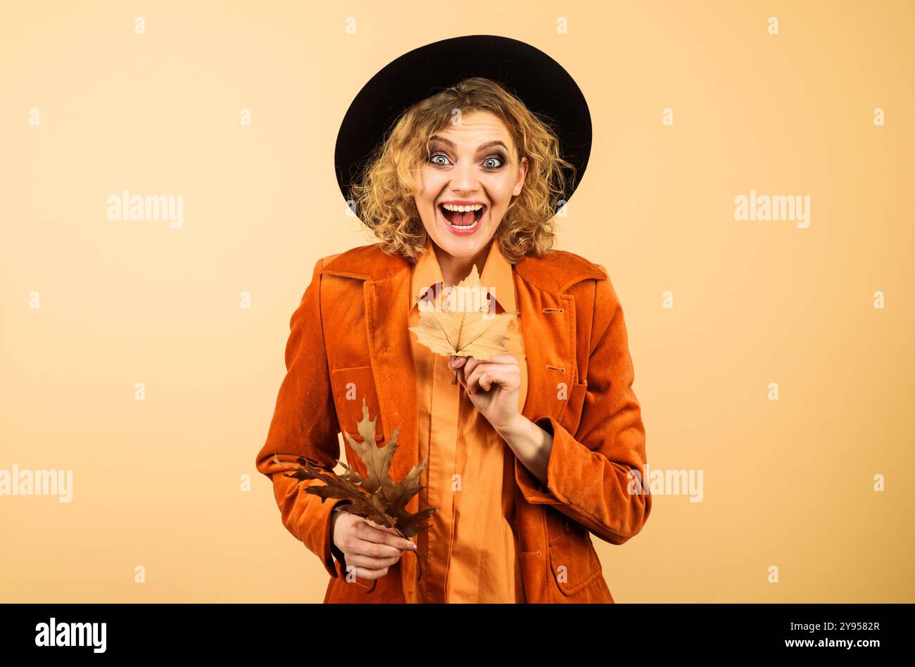 Moda autunnale per le donne. Donna felice con foglie autunnali in cappello nero e giacca arancione. Ragazza autunnale alla moda con un elegante abbigliamento autunnale Foto Stock