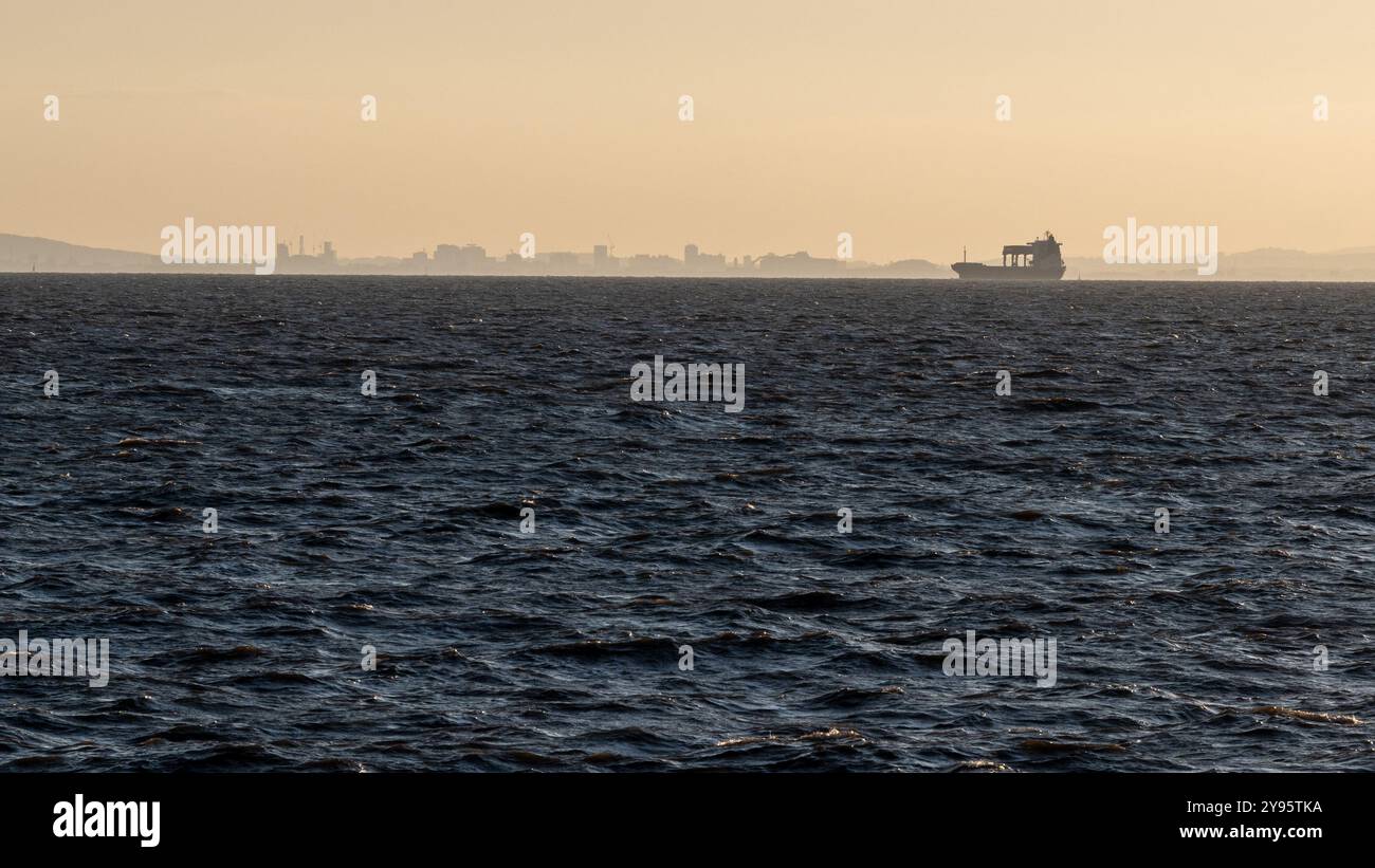 Una nave da carico viaggia lungo il Canale di Bristol al tramonto, con lo skyline di Cardiff, Galles, in sagoma alle spalle. Foto Stock