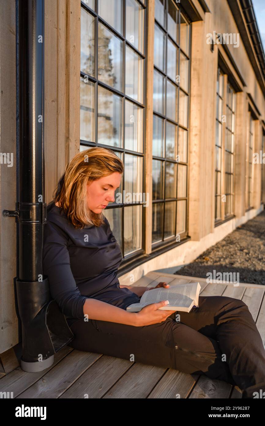 La bella donna siede pacificamente su un ponte di legno, avvolto in un libro mentre la calda luce del sole che tramonta esalta l'atmosfera serena intorno a lei Foto Stock