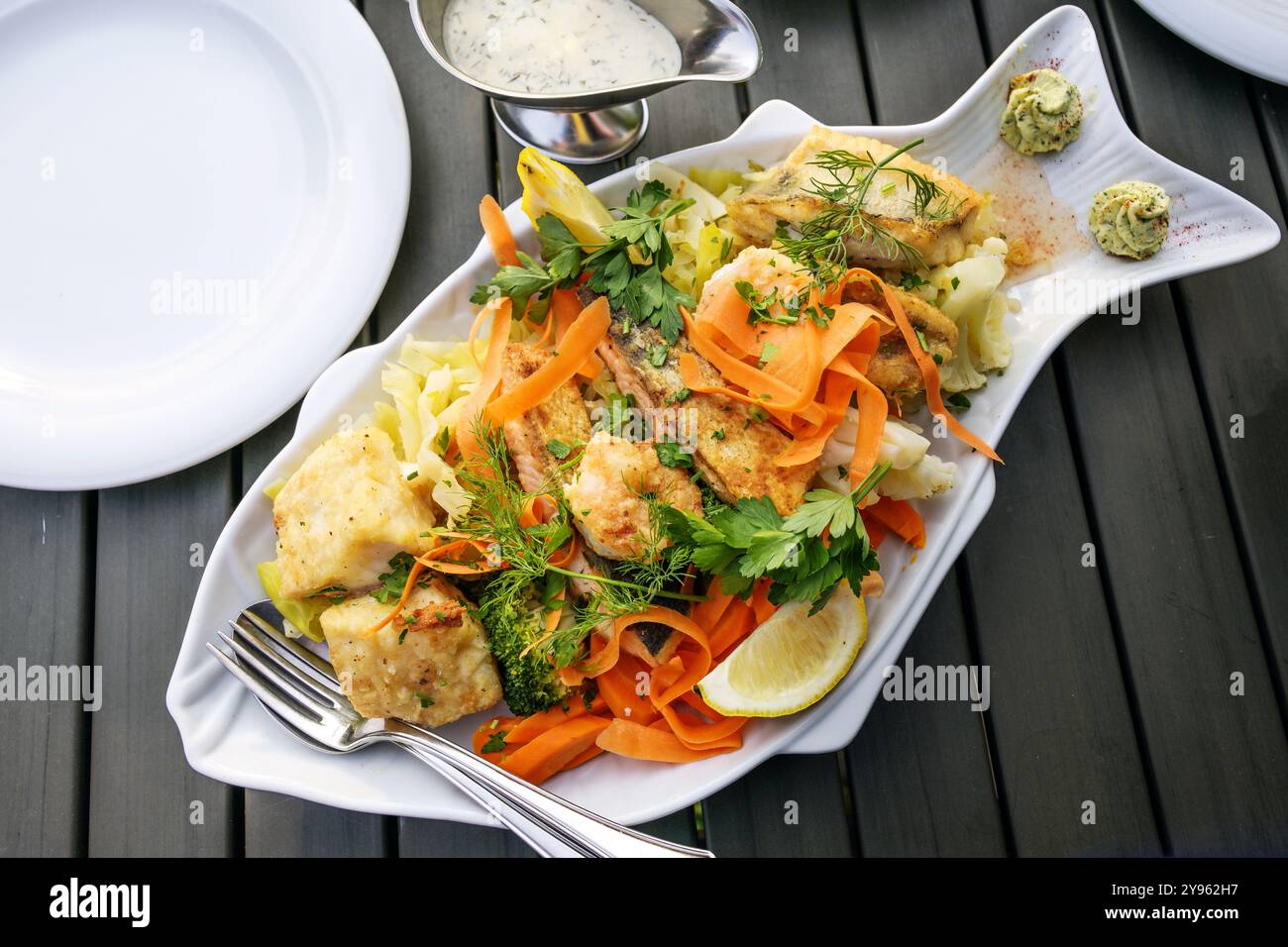 Ampio piatto di pesce con vari filetti di pesce fritti, verdure, limone, erbe e salsa, servito con piatti bianchi su un tavolo di legno scuro, visto da abo Foto Stock