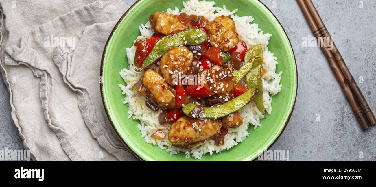 Pollo asiatico agrodolce e appiccicoso con verdure, friggere e riso in una ciotola di ceramica con bacchette, vista dall'alto su fondo in pietra rustica grigia, mestiere Foto Stock