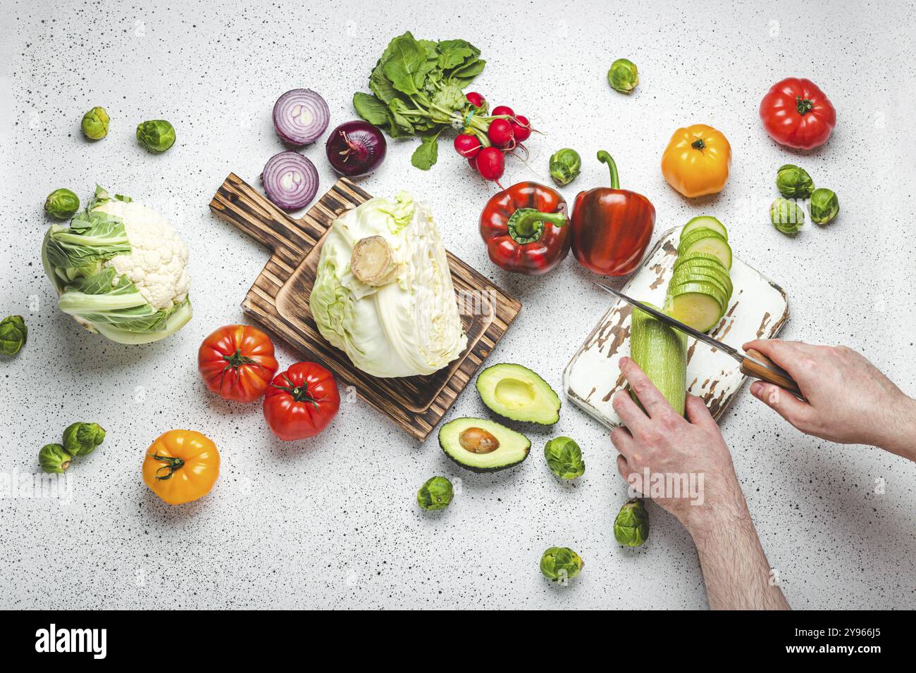 Verdure fresche e mani maschili che tagliano le zucchine con il coltello su tagliere di legno, vista dal tavolo della cucina bianco. Cucina vegetariana da Heal Foto Stock