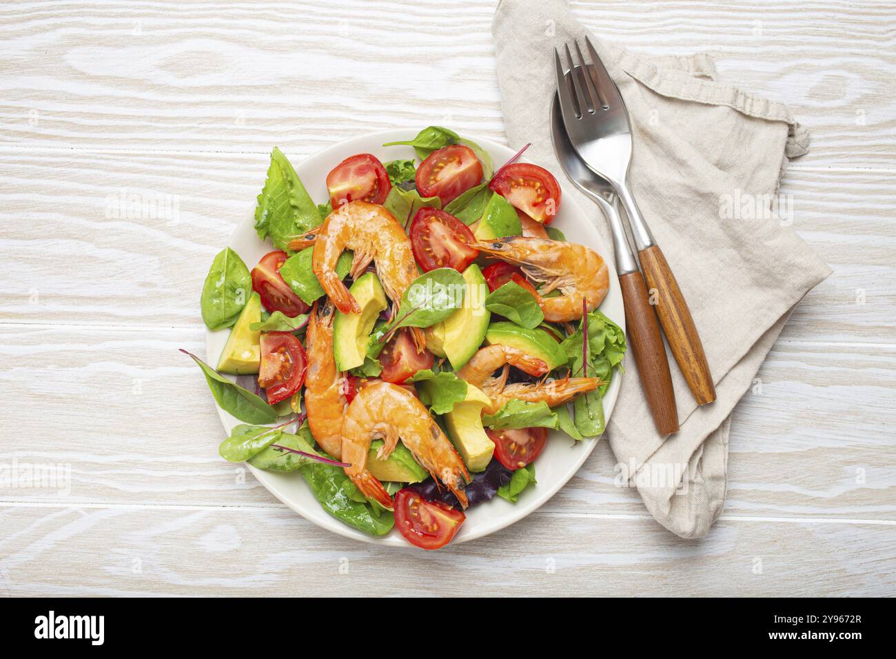 Insalata sana con gamberetti alla griglia, avocado, pomodori ciliegini e foglie verdi su piatto bianco con posate su sfondo rustico in legno bianco vista dall'alto. Foto Stock