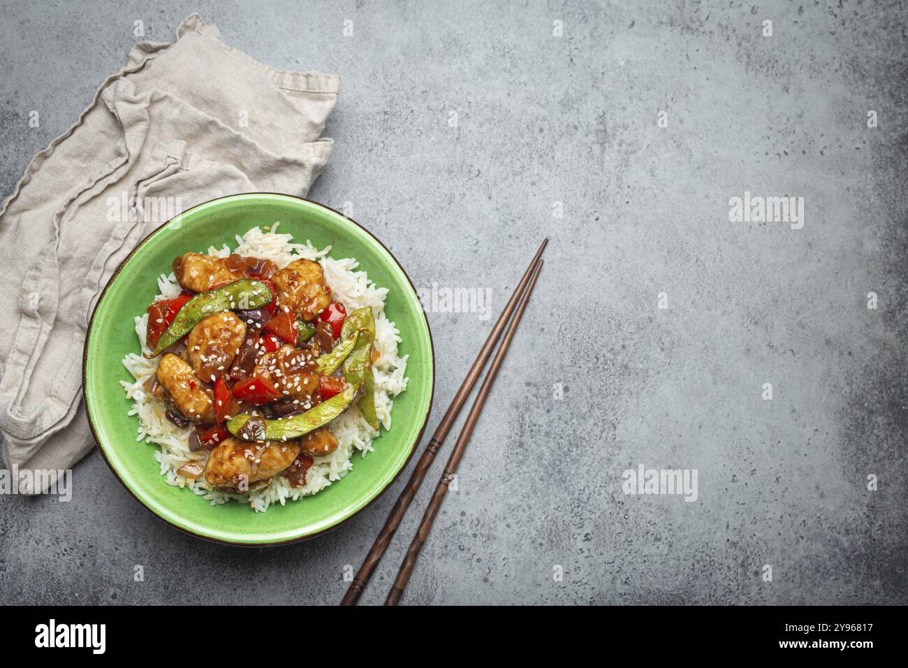 Pollo asiatico agrodolce e appiccicoso con verdure, friggere e riso in una ciotola di ceramica con bacchette, vista dall'alto su fondo in pietra rustica grigia, mestiere Foto Stock