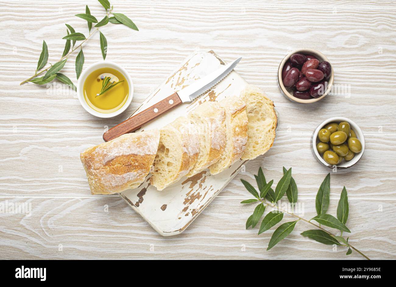 Ciabatta fresca tagliata su tagliere da cucina, olive verdi e marroni, olio d'oliva con rosmarino, rami d'ulivo, fondo rustico in legno bianco t Foto Stock