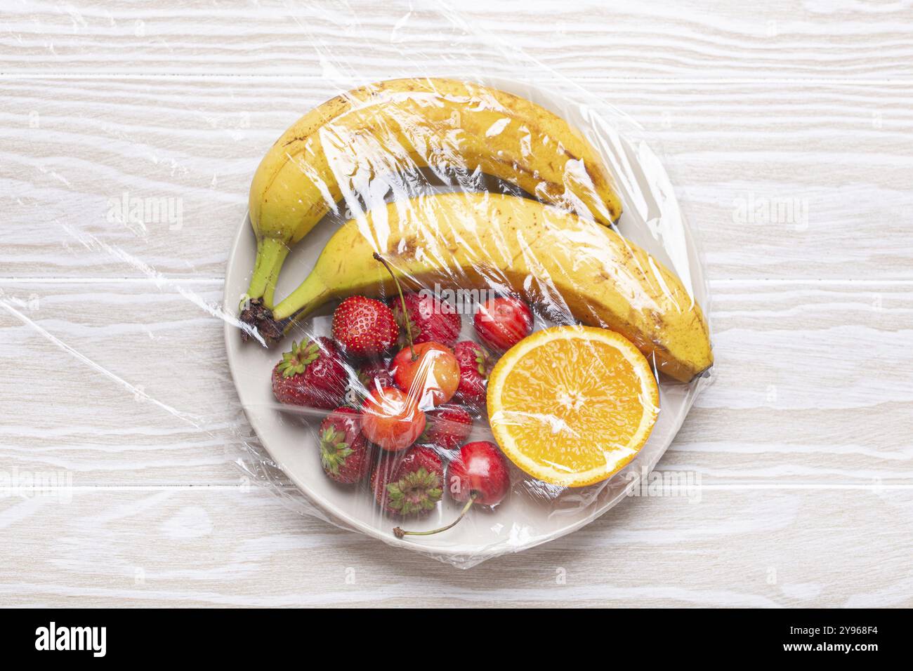 Vari tipi di frutta e frutti di bosco su piatto bianco avvolti dall'alto in pellicola trasparente in plastica su fondo rustico in legno bianco, cibo Foto Stock