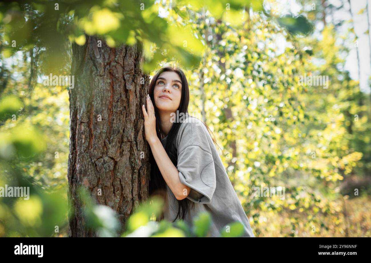 Una donna si appoggia su un albero, guardando verso l'alto con un'espressione serena, circondata da foglie verdi vibranti in un ambiente di foresta illuminato dal sole. Foto Stock