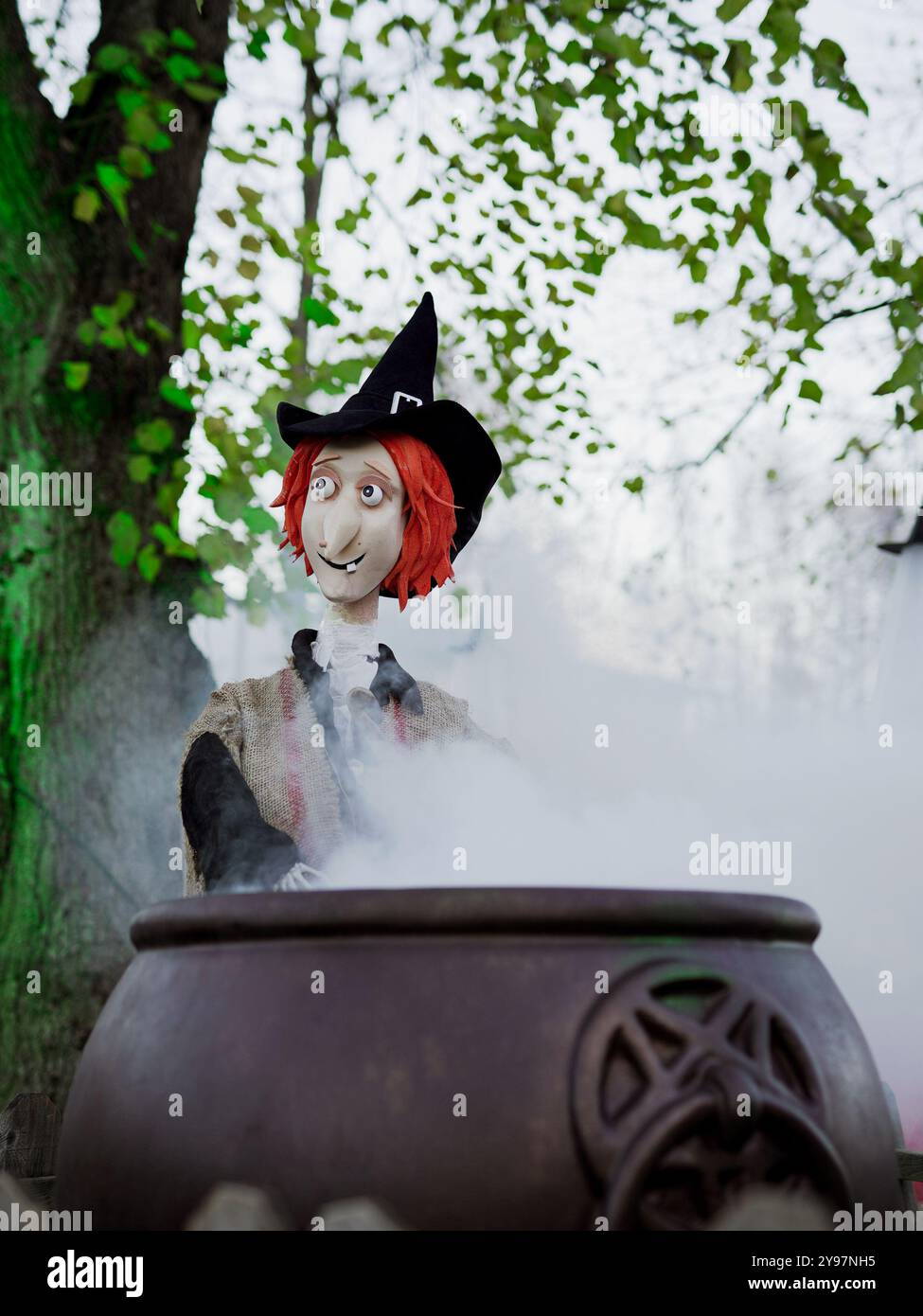 Una stravagante strega di Halloween mescola un calderone fumoso, vestito con un cappello da strega nero e un mantello rustico, che dona un'atmosfera giocosa e spettrale. Foto Stock