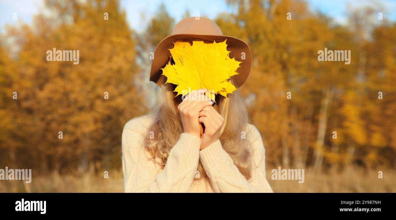 Ritratto di una donna allegra che si copre il viso con foglie d'acero giallo nel parco autunnale, godendosi il caldo sole, la stagione autunnale, il backgroun della natura Foto Stock