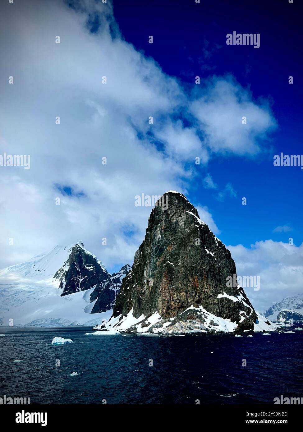 Picco accidentato e innevato che si innalza drammaticamente dalle acque ghiacciate dell'Antartide, contro un cielo blu brillante - Immagine stock catturata con smartphone