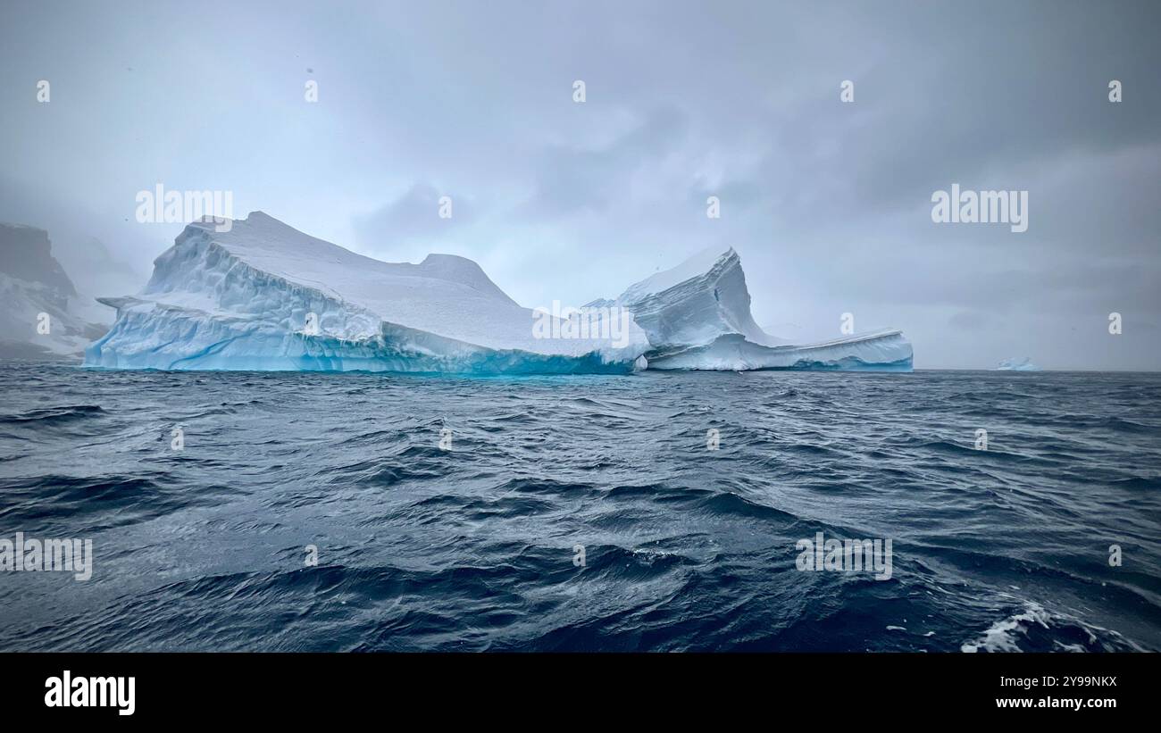 Un enorme iceberg scorre attraverso le acque tempestose dell'Oceano meridionale, con le sue vette frastagliate che si innalzano drammaticamente contro il grigio cielo antartico - Immagine stock catturata con smartphone