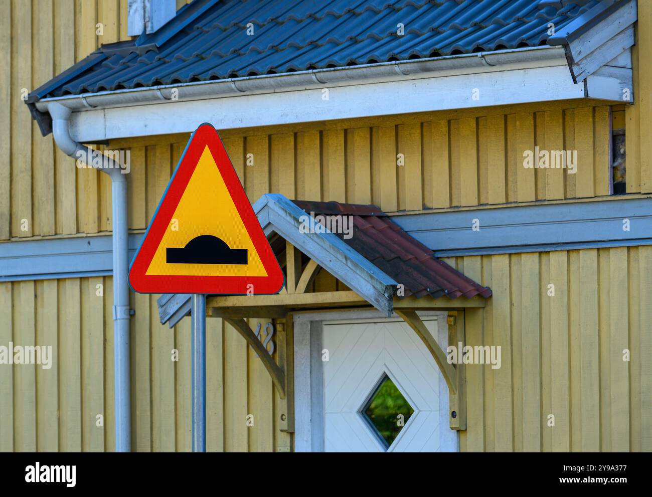 Un segnale luminoso avvisa i conducenti di un dissuasore di velocità, situato vicino a una pittoresca casa di legno gialla con tetto inclinato e portico. La scena è chiara Foto Stock