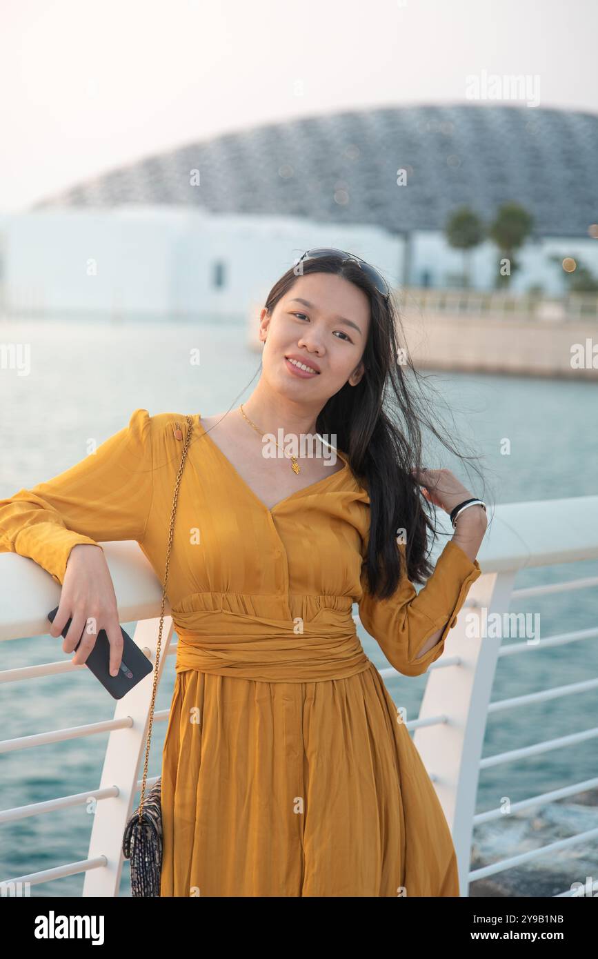 La donna asiatica si erge graziosamente di fronte all'iconico Louvre Abu Dhabi, mescolando cultura e architettura moderna in un suggestivo ambiente artistico Foto Stock