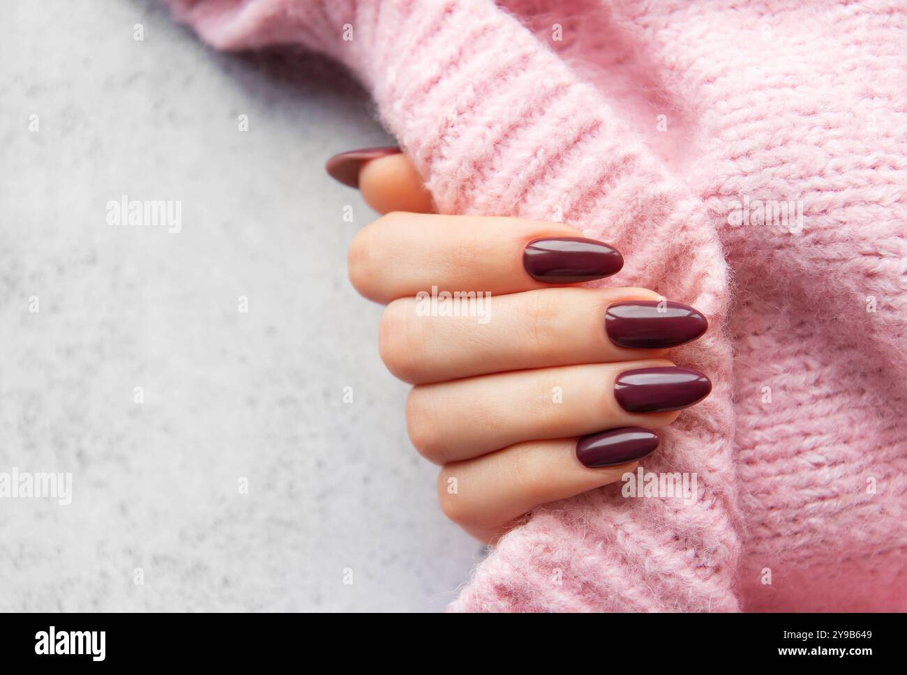 Primo piano della mano di una donna con chiodi a forma di mandorla Bordeaux delicatamente avvolti intorno a un comodo maglione rosa, su uno sfondo neutro Foto Stock