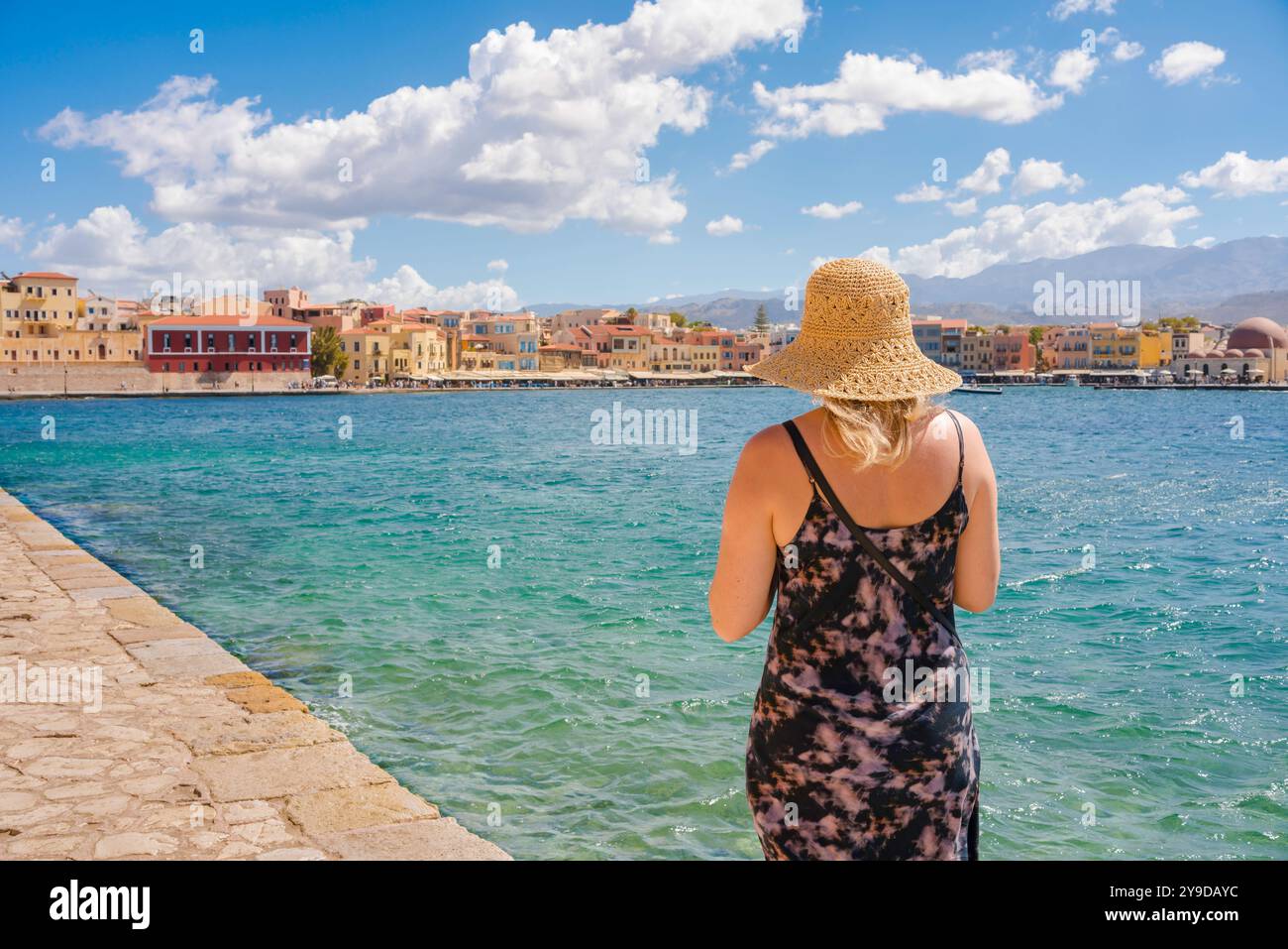 Vacanze estive, vista posteriore di una giovane donna bionda che indossa un cappello estivo che guarda attraverso il porto interno di Chania (Hania), a nord-ovest di Creta, in Grecia Foto Stock