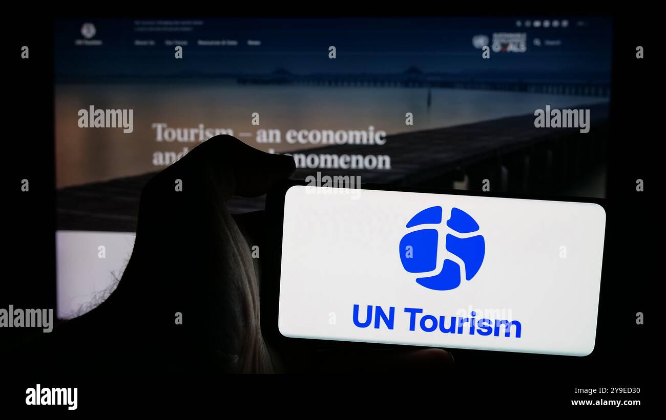 Persona che possiede un telefono cellulare con il logo dell'agenzia delle Nazioni Unite per il turismo delle Nazioni Unite sullo schermo di fronte alla pagina web. Mettere a fuoco il display del telefono. Foto Stock