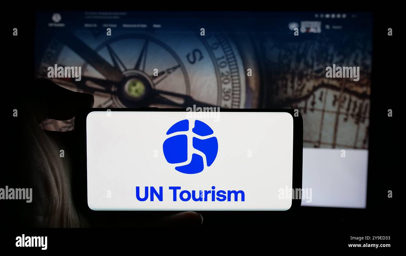Persona che detiene un cellulare con il logo dell'agenzia delle Nazioni Unite per il turismo delle Nazioni Unite sullo schermo di fronte alla pagina web. Mettere a fuoco il display del telefono. Foto Stock