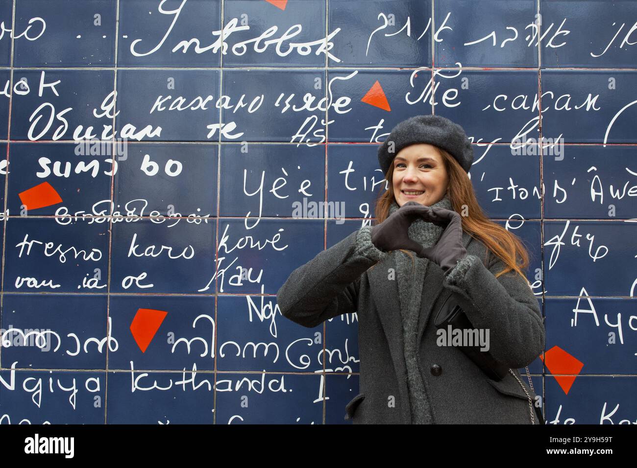 Ragazza in cappotto grigio e berretto vicino al muro d'amore a Parigi mostra cuore Foto Stock