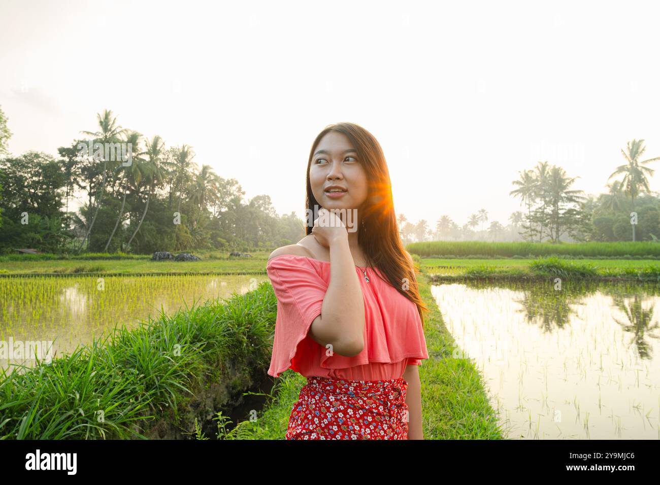Una bella ragazza asiatica si sta tenendo il collo. La ragazza che indossa un vestito rosa. C'è un'esplosione di sole sullo sfondo. Foto Stock