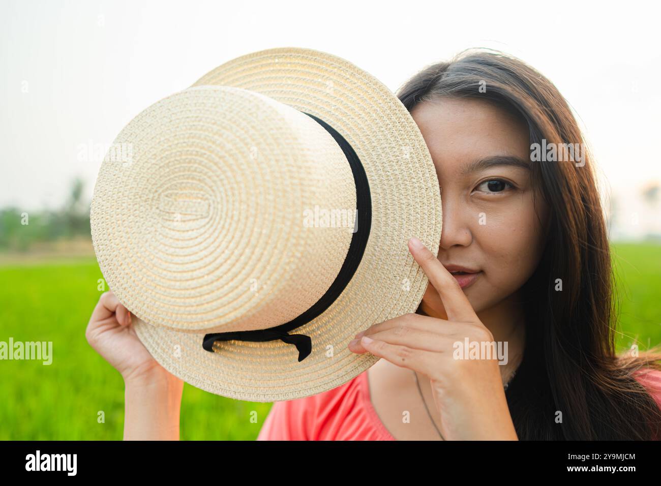 Una bella ragazza asiatica regge un cappello in rattan con l'occhio sinistro. La ragazza che indossa un vestito rosa. Foto Stock