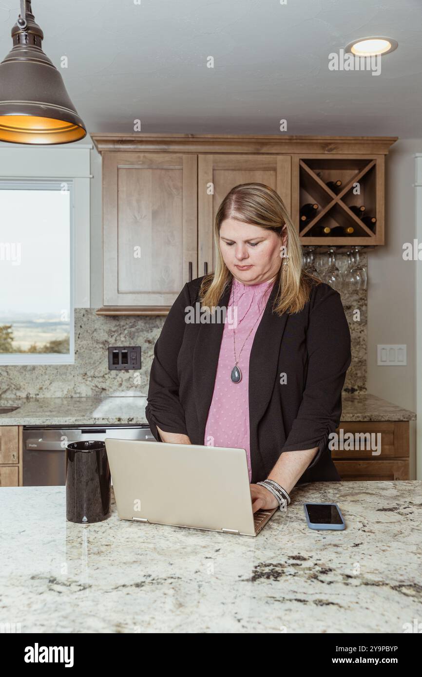 Donna che lavora su un notebook al bancone della cucina con una tazza da caffè Foto Stock