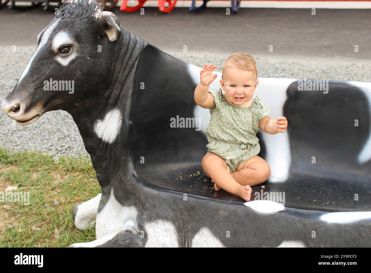 La bambina, in verde, è seduta sulla panchina a forma di mucca durante l'estate Foto Stock