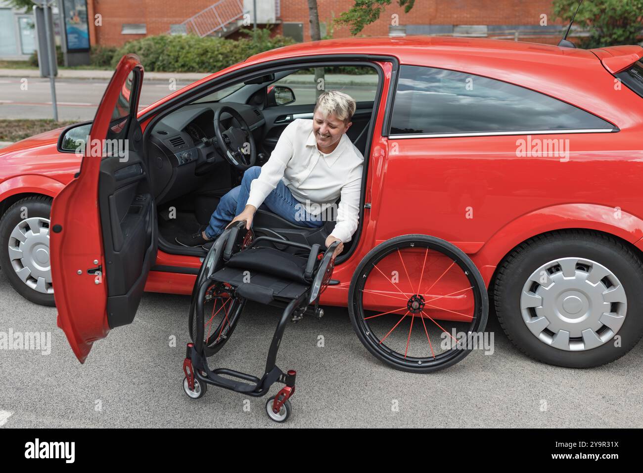 Donna con disabilità che toglie la sedia a rotelle dall'auto mentre siede sul sedile del conducente. Concetto di attrezzature accessibili e adattabili. Foto Stock